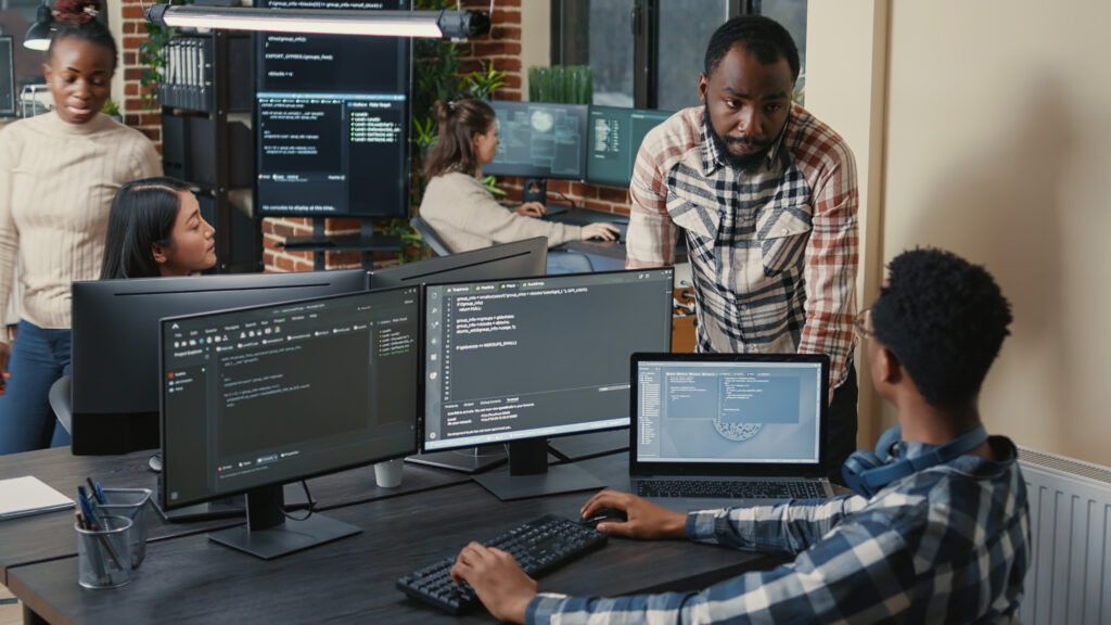 programer sitting desk with multiple screens running code talking with colleague about artificial intelligence algorithm software developers doing innovative artificial intelligence project